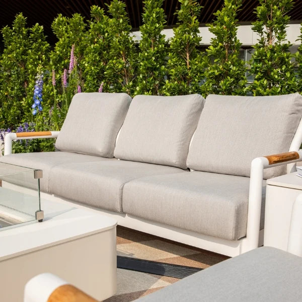 A modern outdoor sofa with light beige cushions and white frame, featuring wooden arm accents, set in a garden patio environment.