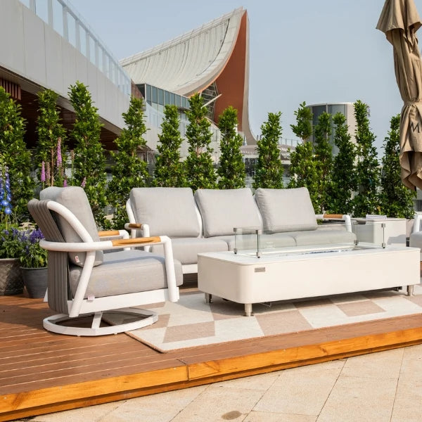Modern outdoor patio furniture set including a white-framed swivel armchair, a three-seat sofa with light gray cushions, a low rectangular coffee table, and a patterned rug on a wooden deck with potted trees and a building in the background.